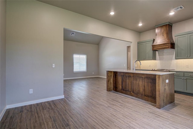 kitchen featuring decorative backsplash, custom exhaust hood, stainless steel gas cooktop, a center island with sink, and light hardwood / wood-style floors