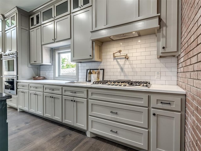 kitchen featuring gray cabinetry, backsplash, dark wood-type flooring, stainless steel appliances, and brick wall