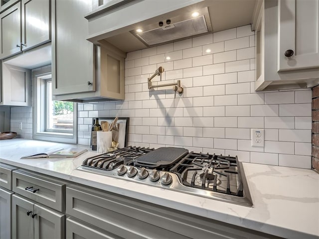 kitchen with light stone counters, ventilation hood, stainless steel gas stovetop, gray cabinets, and decorative backsplash