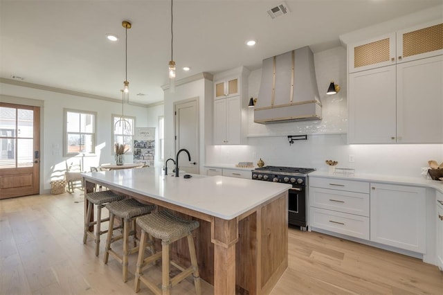 kitchen featuring custom exhaust hood, a kitchen island with sink, light wood-type flooring, stainless steel range, and white cabinetry