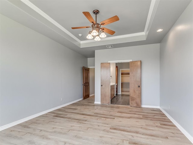 unfurnished bedroom featuring a raised ceiling, ceiling fan, ensuite bathroom, and light wood-type flooring