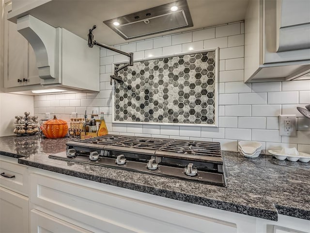 kitchen featuring stainless steel gas cooktop, ventilation hood, backsplash, dark stone countertops, and white cabinets