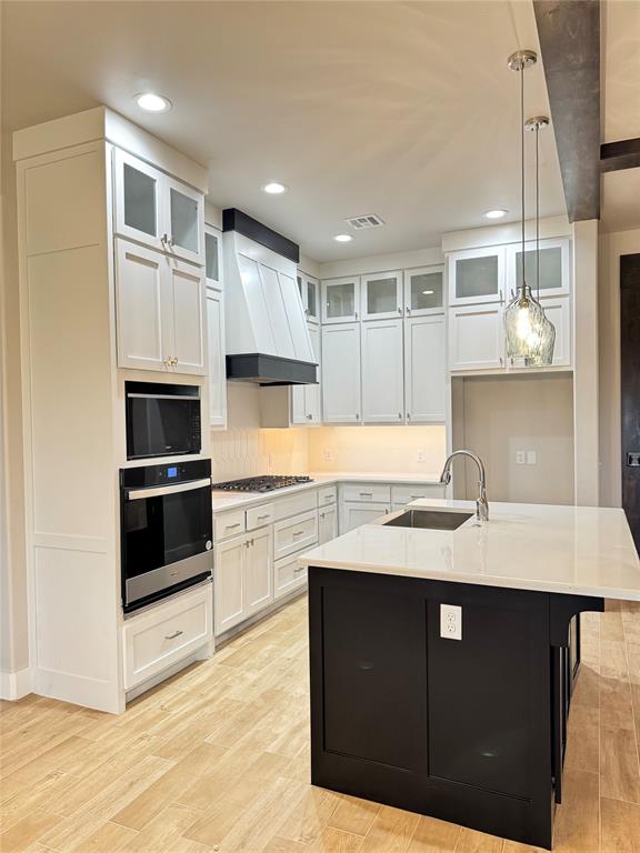 kitchen featuring appliances with stainless steel finishes, white cabinetry, hanging light fixtures, a kitchen island with sink, and custom range hood