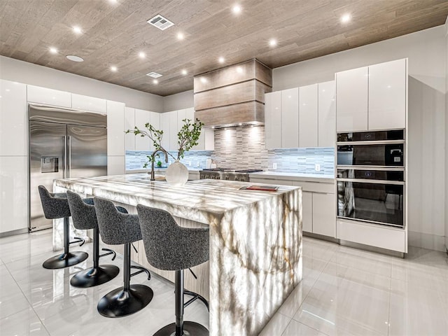 kitchen with white cabinetry, stainless steel appliances, premium range hood, and an island with sink