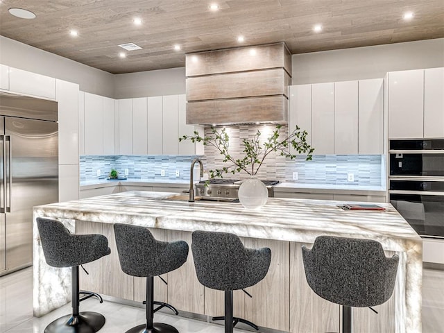 kitchen with a breakfast bar, white cabinetry, stainless steel built in refrigerator, and an island with sink