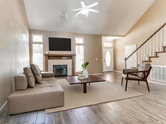 living room featuring high vaulted ceiling, light hardwood / wood-style floors, a tile fireplace, and ceiling fan