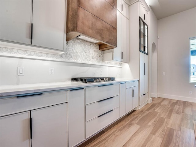 kitchen with backsplash, custom exhaust hood, light hardwood / wood-style floors, white cabinetry, and stainless steel gas stovetop