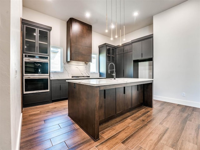 kitchen featuring pendant lighting, a kitchen island with sink, stainless steel double oven, and light hardwood / wood-style floors