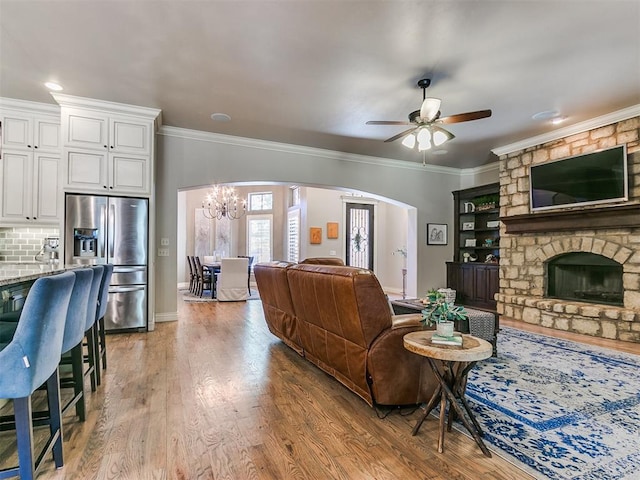 living room with ceiling fan with notable chandelier, light hardwood / wood-style floors, a stone fireplace, and crown molding