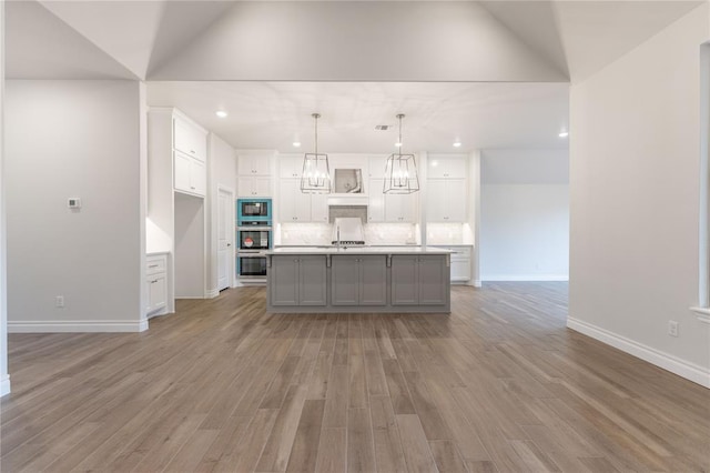 kitchen with decorative light fixtures, white cabinetry, a kitchen island with sink, stainless steel double oven, and light hardwood / wood-style flooring