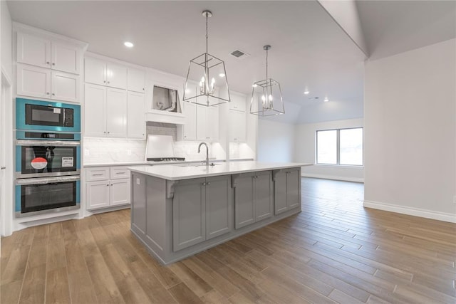 kitchen with white cabinetry, backsplash, hanging light fixtures, a kitchen island with sink, and light wood-type flooring