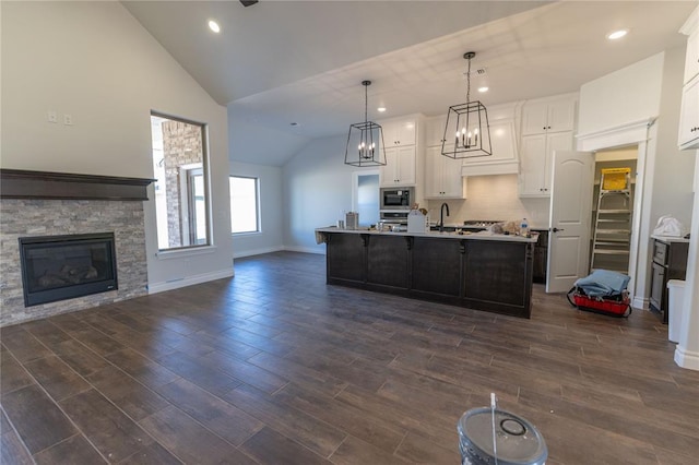 kitchen with a center island with sink, white cabinetry, hanging light fixtures, built in microwave, and a stone fireplace