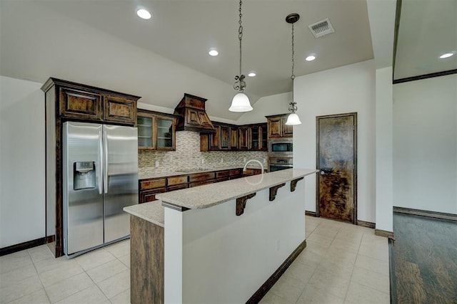 kitchen featuring premium range hood, light stone countertops, an island with sink, appliances with stainless steel finishes, and dark brown cabinets