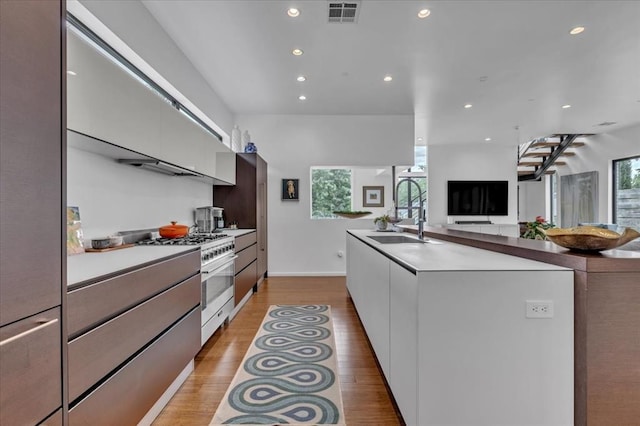 kitchen featuring a kitchen island with sink, sink, light wood-type flooring, stainless steel range, and white cabinetry