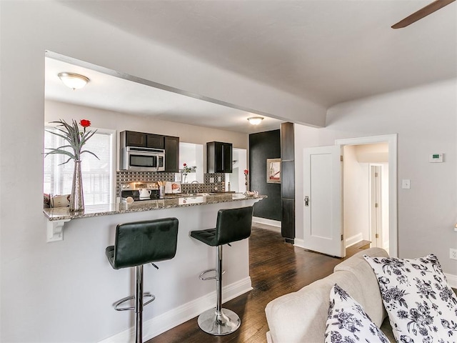 kitchen with decorative backsplash, appliances with stainless steel finishes, light stone countertops, a breakfast bar, and dark wood-type flooring