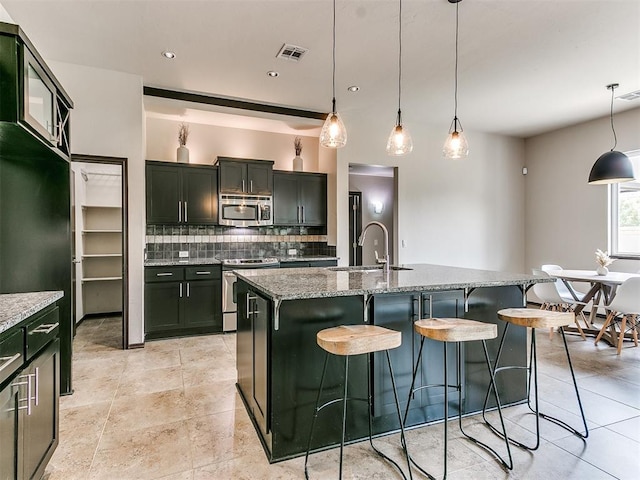 kitchen featuring stainless steel appliances, light stone counters, hanging light fixtures, and a center island with sink