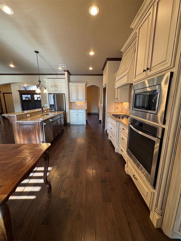 kitchen with light stone counters, a center island with sink, dark wood finished floors, arched walkways, and stainless steel appliances