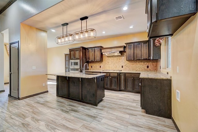 kitchen with light stone countertops, dark brown cabinets, stainless steel appliances, hanging light fixtures, and an island with sink