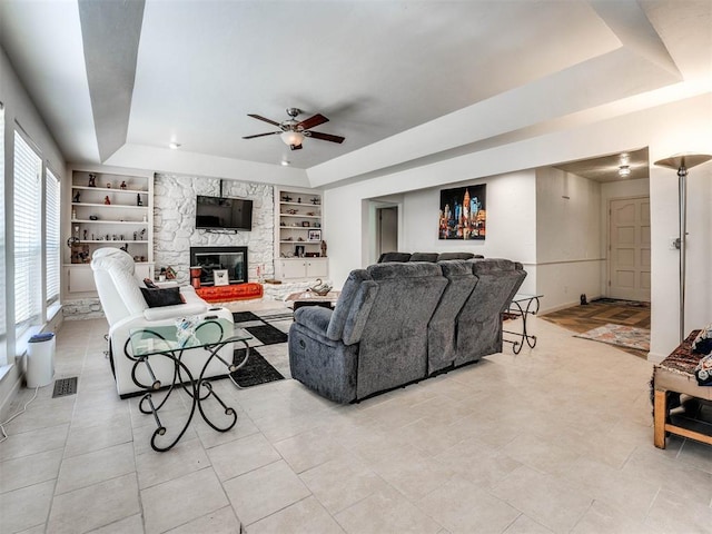 living room with a tray ceiling, built in features, ceiling fan, and a stone fireplace