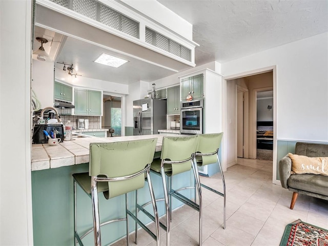 kitchen with tile counters, kitchen peninsula, stainless steel appliances, and green cabinetry