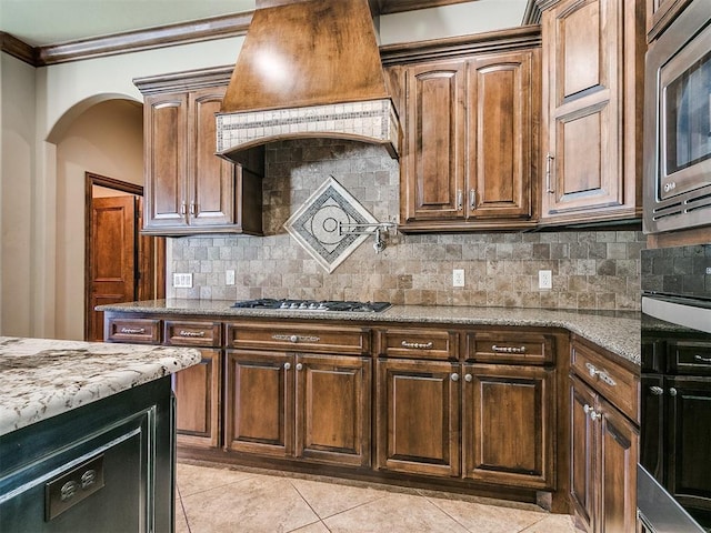 kitchen featuring light stone countertops, custom range hood, light tile patterned floors, decorative backsplash, and appliances with stainless steel finishes