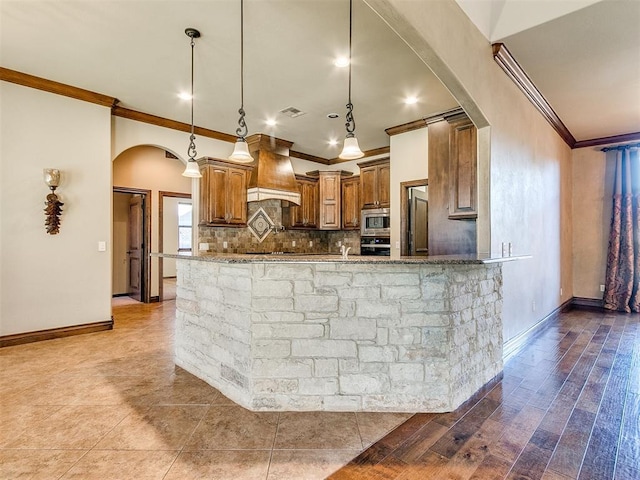 kitchen featuring custom range hood, stainless steel microwave, wall oven, arched walkways, and light stone countertops