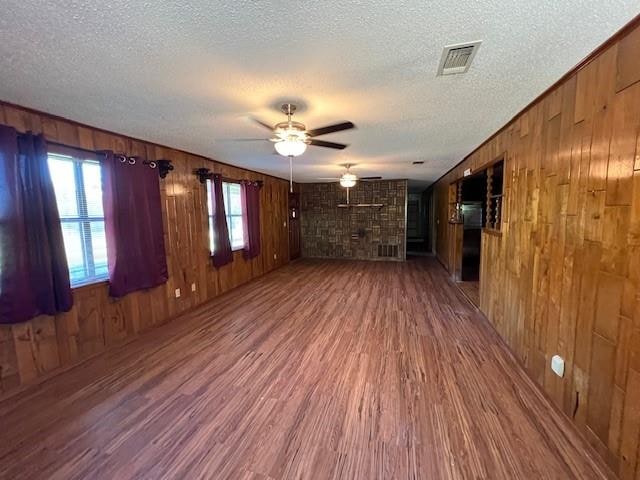 unfurnished living room with a textured ceiling, ceiling fan, hardwood / wood-style flooring, a stone fireplace, and wood walls