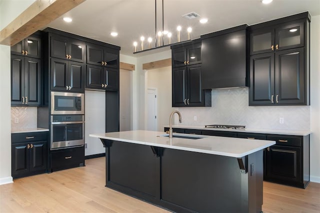 kitchen with sink, stainless steel appliances, a center island with sink, custom exhaust hood, and light wood-type flooring