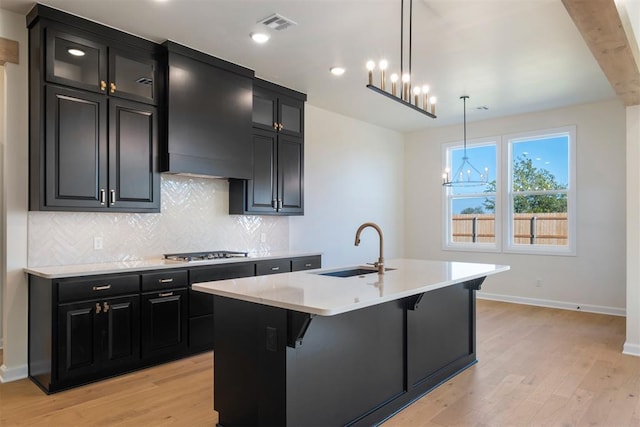 kitchen with light wood-type flooring, custom exhaust hood, sink, pendant lighting, and a center island with sink