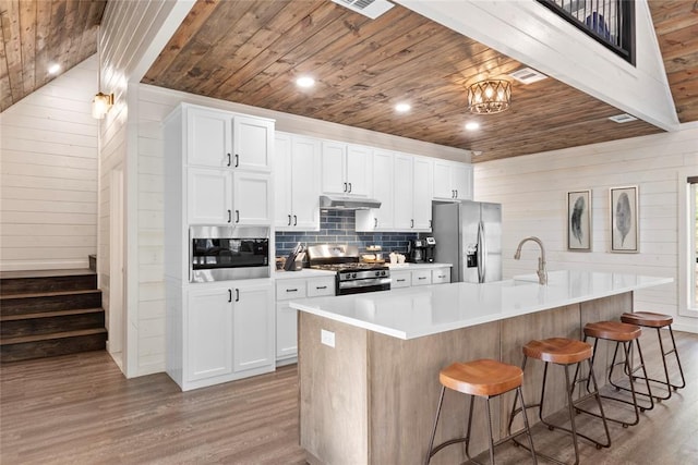 kitchen featuring stainless steel appliances, wooden walls, a large island with sink, hardwood / wood-style flooring, and white cabinets