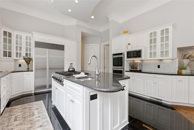 kitchen featuring built in appliances, a kitchen island with sink, white cabinets, and dark stone counters