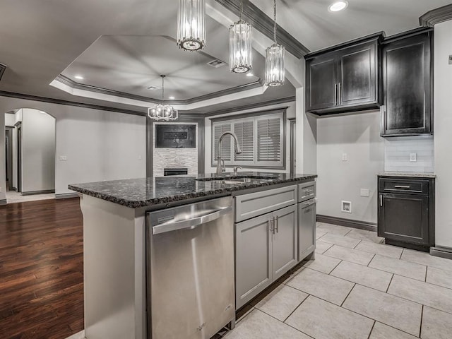 kitchen with dishwasher, a center island with sink, a tray ceiling, and dark stone countertops