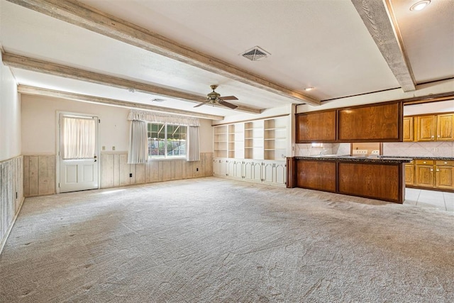 unfurnished living room featuring wood walls, beamed ceiling, light colored carpet, and ceiling fan