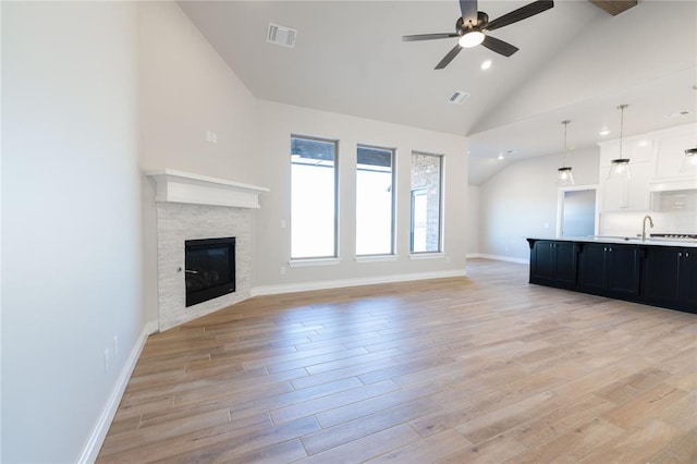 unfurnished living room featuring ceiling fan, sink, a tiled fireplace, light wood-type flooring, and high vaulted ceiling