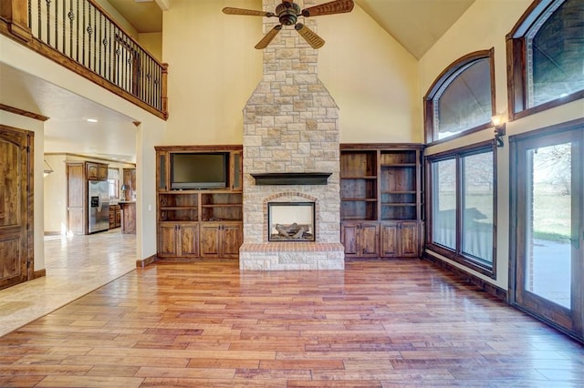 unfurnished living room with high vaulted ceiling, a stone fireplace, light hardwood / wood-style flooring, ceiling fan, and built in features