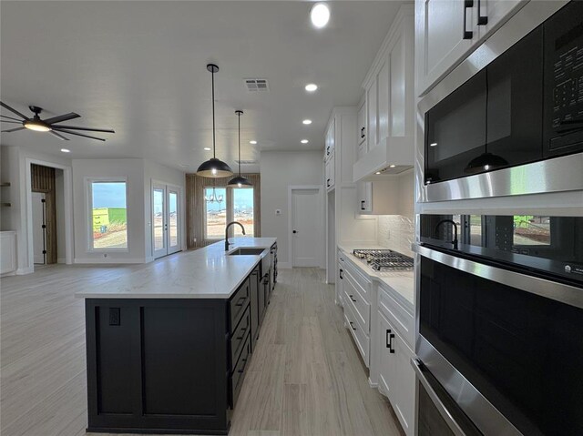 kitchen with appliances with stainless steel finishes, light wood-type flooring, a center island with sink, white cabinetry, and hanging light fixtures