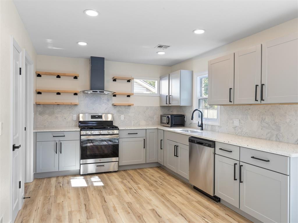 kitchen with gray cabinetry, wall chimney exhaust hood, light wood-type flooring, and appliances with stainless steel finishes