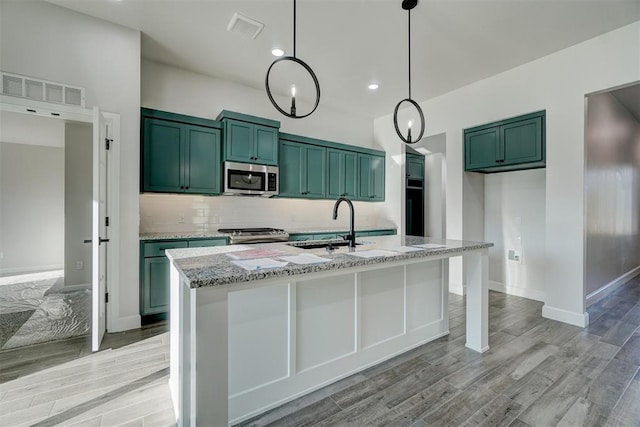 kitchen with light stone countertops, stainless steel appliances, light hardwood / wood-style flooring, hanging light fixtures, and an island with sink