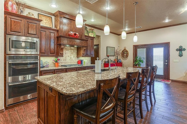 kitchen with stainless steel appliances, a center island with sink, hanging light fixtures, and dark wood-type flooring