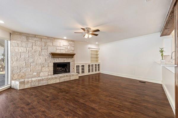 unfurnished living room featuring built in shelves, a stone fireplace, ornamental molding, dark hardwood / wood-style flooring, and ceiling fan