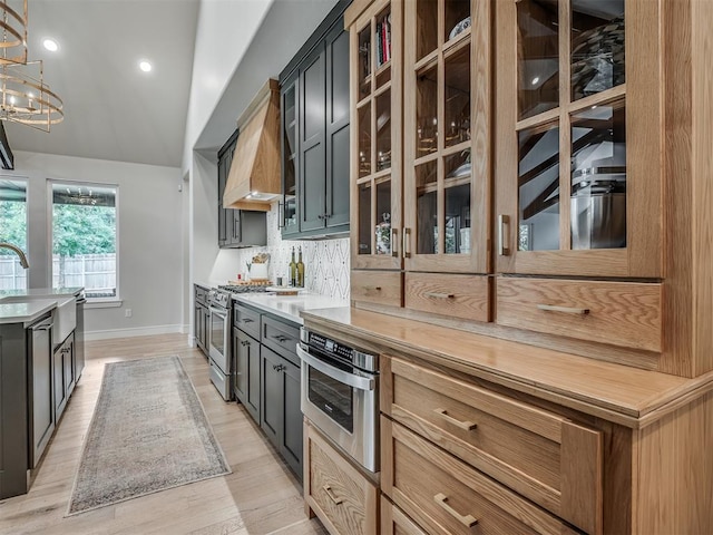 kitchen featuring appliances with stainless steel finishes, backsplash, premium range hood, decorative light fixtures, and light hardwood / wood-style flooring