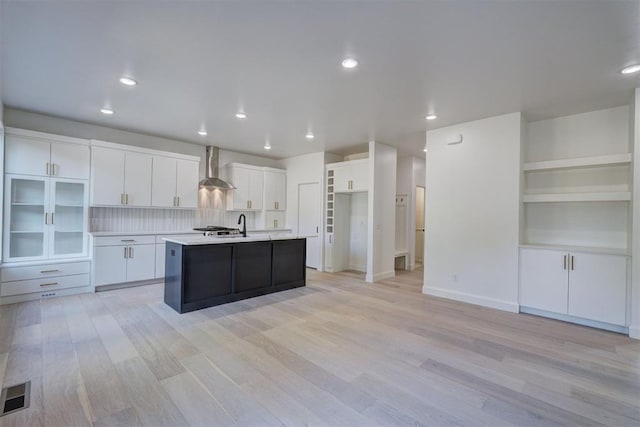 kitchen featuring light countertops, a center island with sink, wall chimney range hood, and white cabinetry