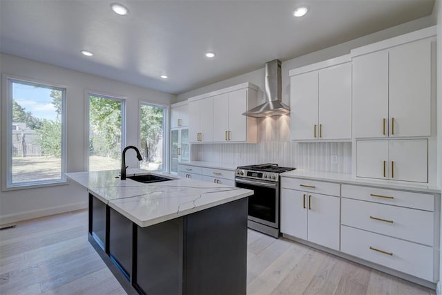 kitchen with stainless steel range with gas cooktop, white cabinets, a kitchen island with sink, a sink, and wall chimney exhaust hood