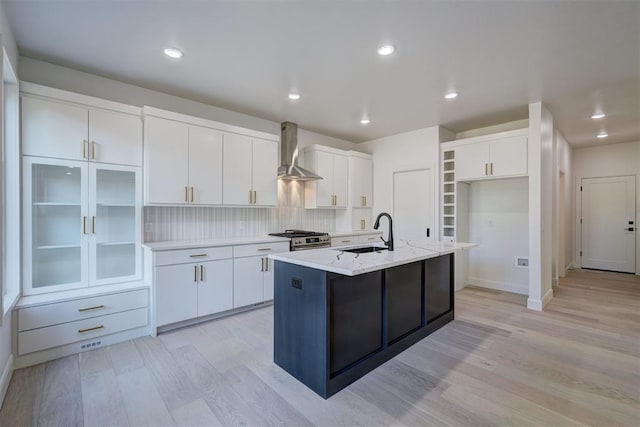 kitchen with a sink, a kitchen island with sink, wall chimney range hood, white cabinetry, and stainless steel range with gas stovetop