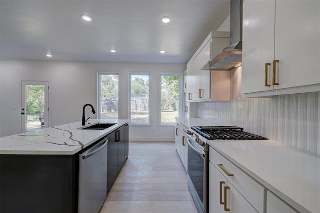 kitchen featuring appliances with stainless steel finishes, a kitchen island with sink, a sink, wall chimney range hood, and white cabinetry