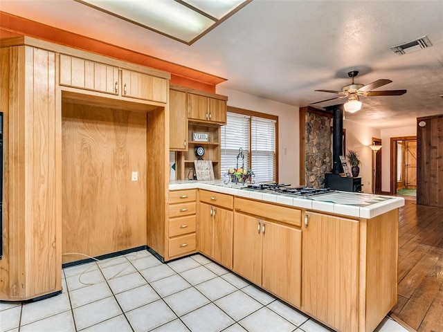 kitchen with tile countertops, a wood stove, ceiling fan, light hardwood / wood-style floors, and stainless steel gas cooktop