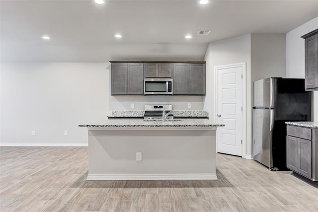 kitchen with light stone countertops, appliances with stainless steel finishes, light wood-type flooring, sink, and a center island with sink