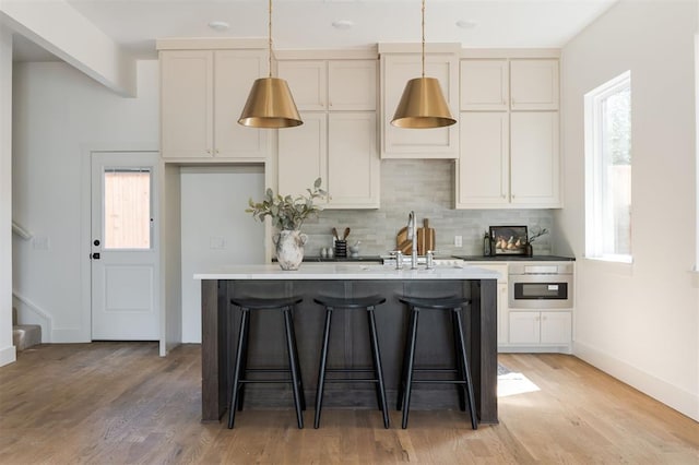 kitchen featuring a kitchen island with sink, oven, light hardwood / wood-style flooring, decorative backsplash, and decorative light fixtures