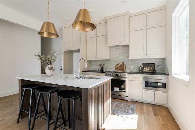 kitchen with a kitchen island with sink, sink, light hardwood / wood-style flooring, appliances with stainless steel finishes, and decorative light fixtures