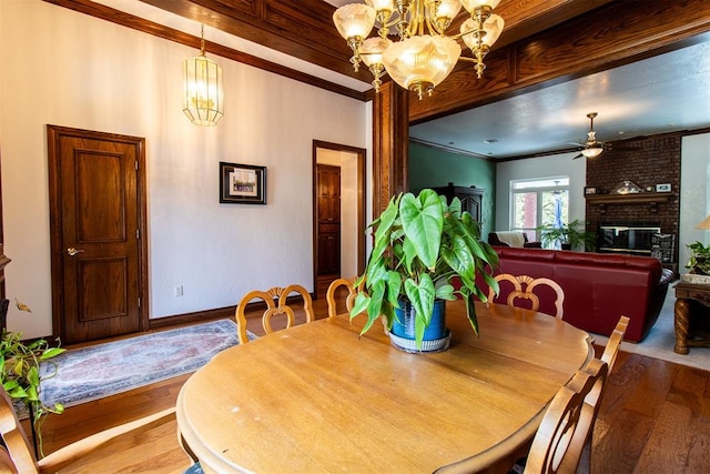 dining space with ceiling fan with notable chandelier, wood-type flooring, crown molding, and a brick fireplace